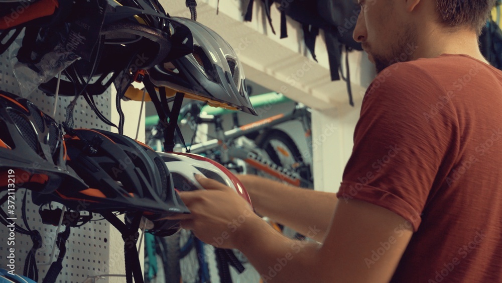 A young Caucasian man stands near a stand in a bicycle store in the head protection department. Choosing a bicycle helmet in a small store A young Caucasian man stands near a stand in a bicycle store in the head protection department. Choosing a bicycle helmet in a small store