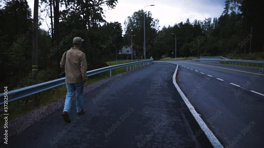 Follow shot of young man with camera walking by side of country road Follow shot of young man with camera walking by side of country road