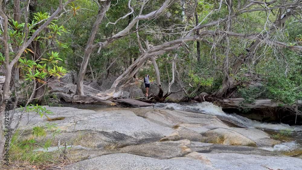 Adult Male Carefully Walking Barefoot Across Rocks And Bridge At Emerald Falls Creek. Locked Off Adult Male Carefully Walking Barefoot Across Rocks And Bridge At Emerald Falls Creek. Locked Off