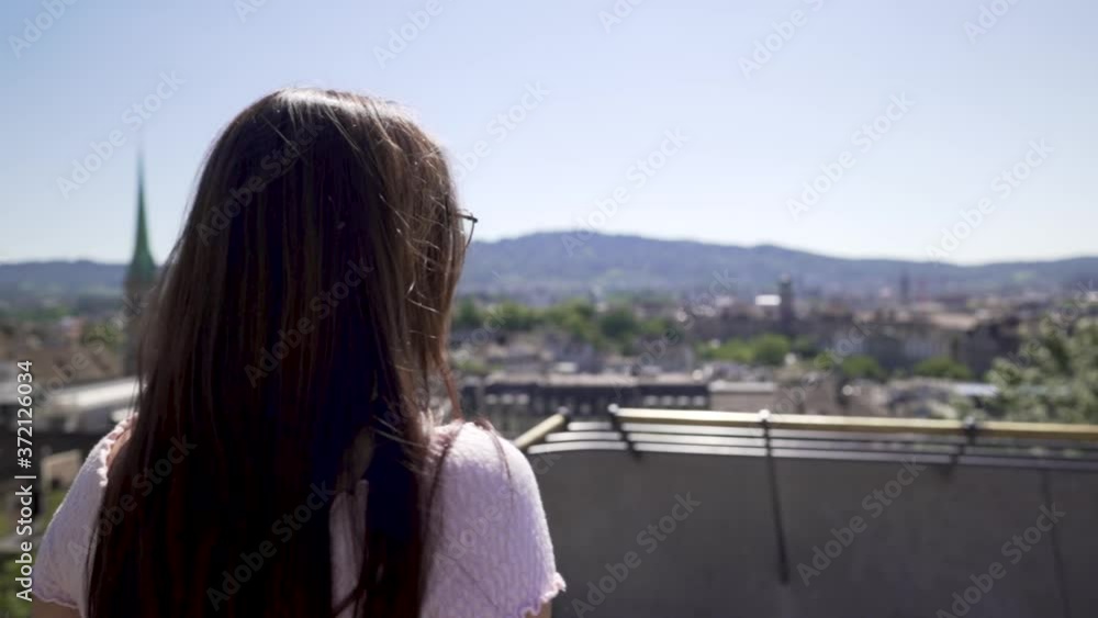 Overshoulder and slow motion shot of young female student in grey jeans and pink top wearing glasses on a terrace over Zurich city smiles in camera Overshoulder and slow motion shot of young female student in grey jeans and pink top wearing glasses on a terrace over Zurich city smiles in camera