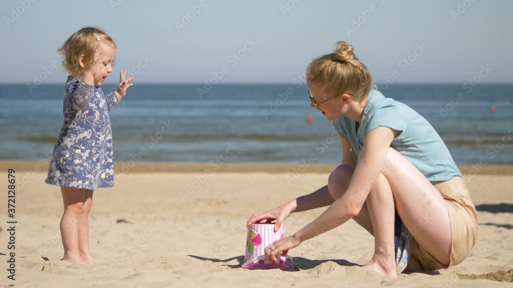 Young beautiful mother tries to show her daughter how to built sand castle. Family vacation near the sea. Mother and daughter wear similar color clothes. Young beautiful mother tries to show her daughter how to built sand castle. Family vacation near the sea. Mother and daughter wear similar color clothes.