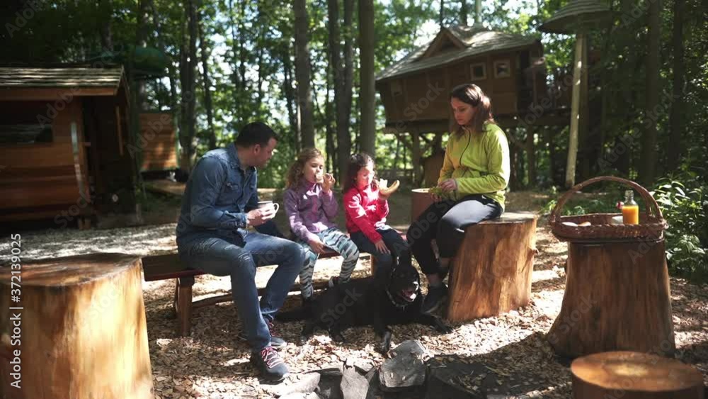 Static FHD shot of a family with two daughters sitting on a bench in a forest in Dolní Morava, Czechia, having a snack and enjoying the day. Family sitting at a forest campsite and having a snack. Static FHD shot of a family with two daughters sitting on a bench in a forest in Dolní Morava, Czechia, having a snack and enjoying the day. Family sitting at a forest campsite and having a snack.