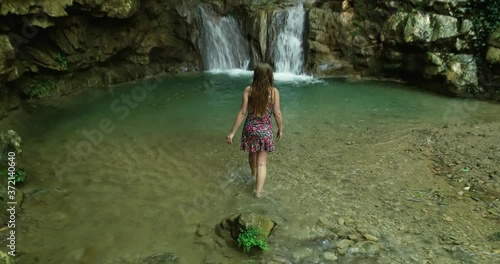 Scenic landscape view above young Caucasian woman sitting on rock in shallow water, stands up and walks towards clear fresh Yahshoush mountain waterfall, Lebanon, behind overhead drone pull back photo