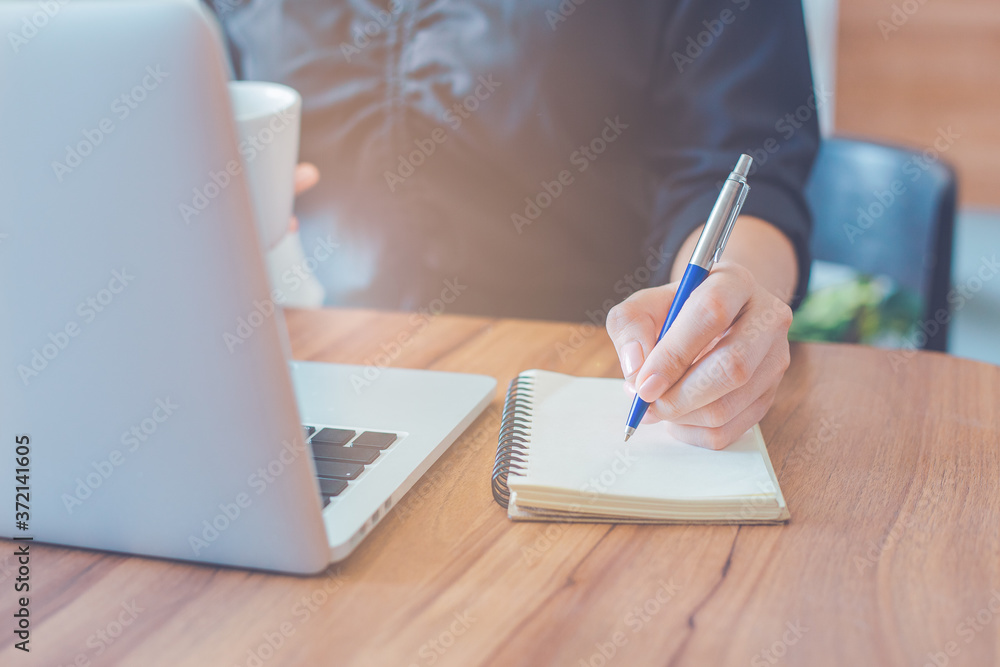 Woman is writing on a notebook with a pen and using a laptop to work in the office.