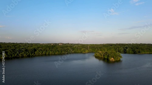 Drone circling around small green island in the middle of the deep blue lake surrounded by thick forest vegetation. Aerial view of Hyland Lake Park in Bloomington, Minnesota. photo