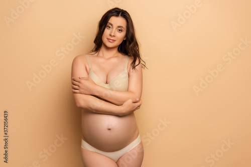 pregnant woman in lingerie standing with crossed arms while looking at camera on beige