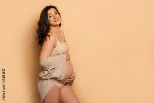  pregnant woman in lingerie touching tummy and looking at camera on beige