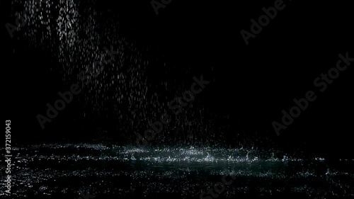 Spray spatters rain fall against on the water surface which glow. Black background. White neon lighting in dark studio. Slow motion.