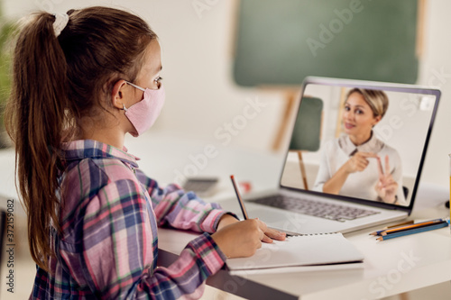 Schoolgirl writing while having online class with her teacher over laptop due to virus pandemic.