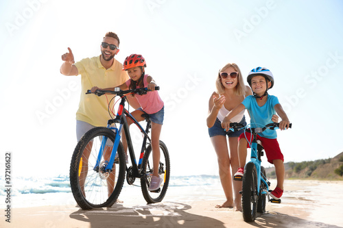 Happy parents teaching children to ride bicycles on sandy beach near sea