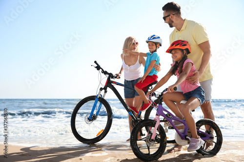 Happy parents teaching children to ride bicycles on sandy beach near sea