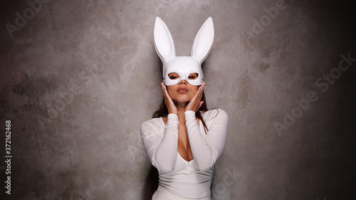 Portrait of beautiful flirting Asian woman in white dress and rabbit mask playfully posing for camera in the spotlight