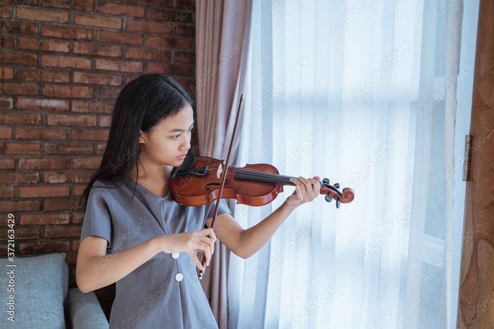 teenage girl learning to play a violin instrument in a room at home against a curtain background teenage girl learning to play a violin instrument in a room at home against a curtain background
