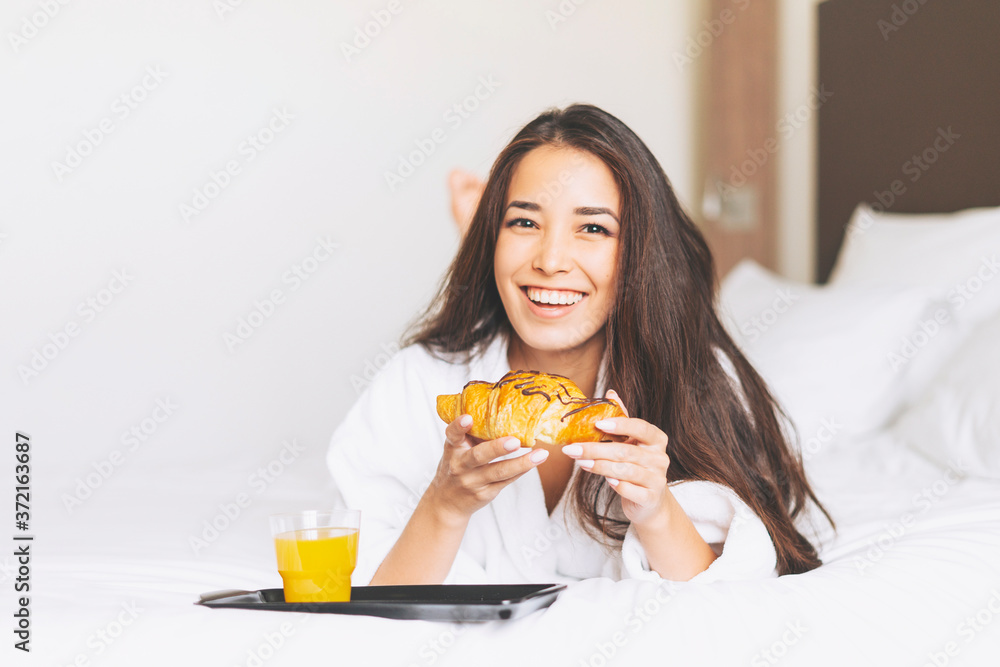 Beautiful happy Asian girl with long hair in white robe having breakfast Croissant and orange juice in the bed of hotel room Beautiful happy Asian girl with long hair in white robe having breakfast Croissant and orange juice in the bed of hotel room