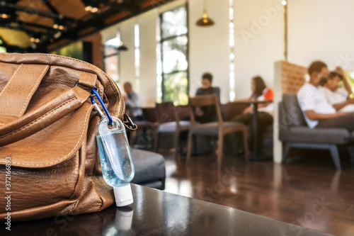 Mini portable alcohol gel bottle to kill Corona Virus(Covid-19) hang on a brown leather shoulder bag on table in coffee shop.New normal lifestyle. Health care concept. Selective focus on alcohol gel
