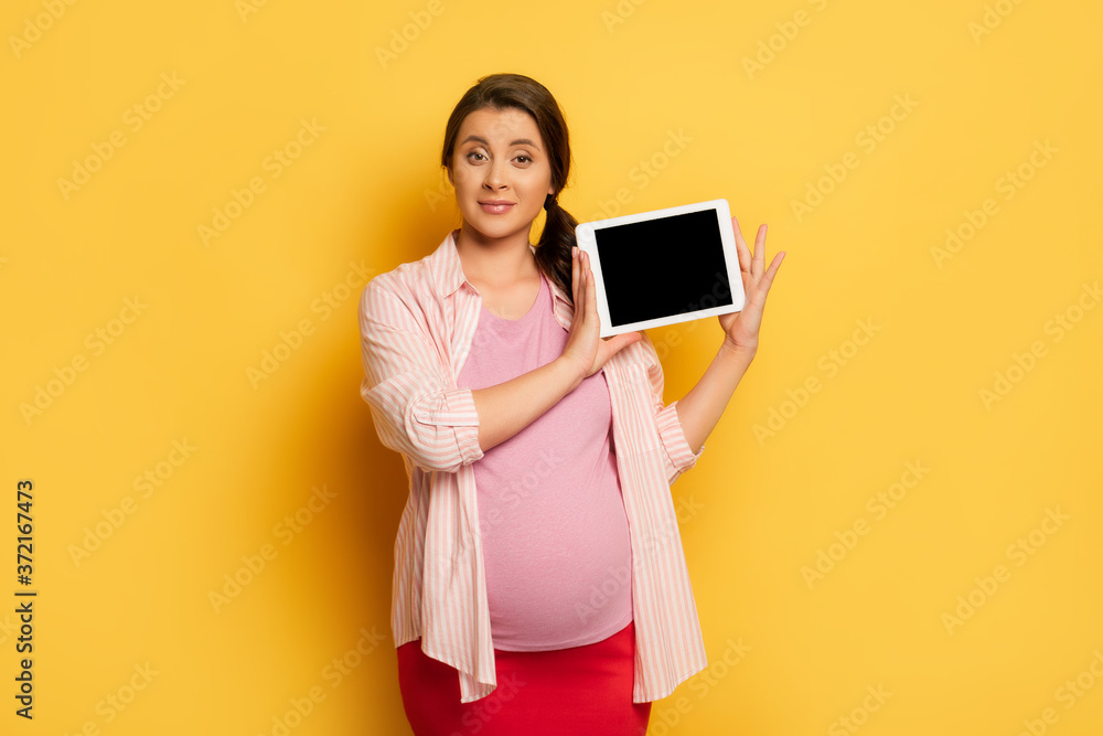 pregnant woman looking at camera while showing digital tablet with blank screen on yellow background pregnant woman looking at camera while showing digital tablet with blank screen on yellow background
