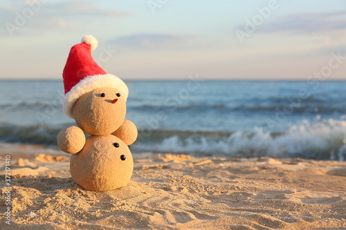 Snowman made of sand with Santa hat on beach near sea  space for text. Christmas vacation