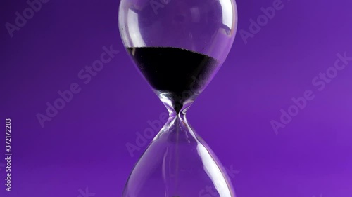 Shot of a sand clock measuring time while the sand is falling down against the purple background - old classic timer. Extreme close up of a transparent hourglass with flowing black sand - time concept