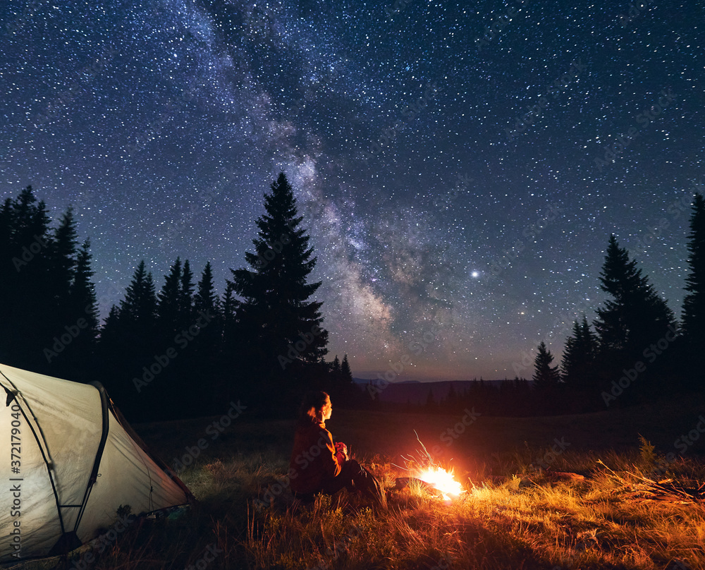Back view of woman sitting by bright burning campfire near tent alone, enjoying beautiful camping night under dark sky full of stars and bright Milky Way, warm summer evening in the mountains. - obrazy, fototapety, plakaty 