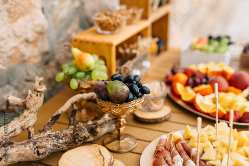 Close-up of a gilded bowl of grapes and figs on a table with sliced ham, cheese and fruit in plates.