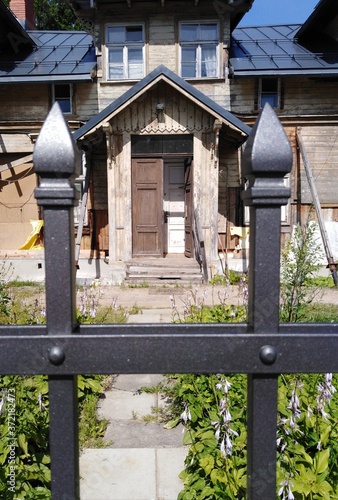 Decorative carved metal fence in front of a wooden old house, which is being restored on a sunny summer day.