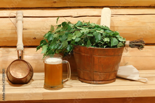 A glass mug of light beer in the interior of sauna among copper vat, ladle and fresh birch broom.
A mug of beer in the sauna.