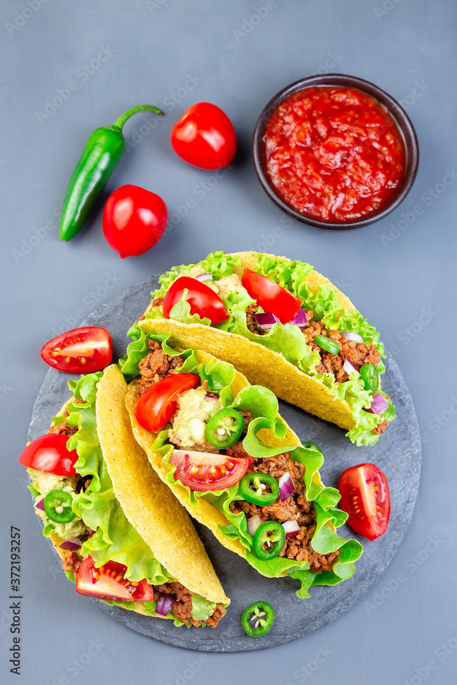 Taco shells on stone plate, with lettuce, ground beef meat, mashed avocado, tomato, red onion and jalapeno pepper, vertical, top view Taco shells on stone plate, with lettuce, ground beef meat, mashed avocado, tomato, red onion and jalapeno pepper, vertical, top view