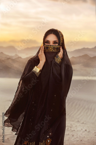 portrait of young beautiful oriental mysterious girl. The woman looks into the camera. The face is hidden by a golden veil. Luxurious evening make-up, smoky eyes. The head is covered with black scarf