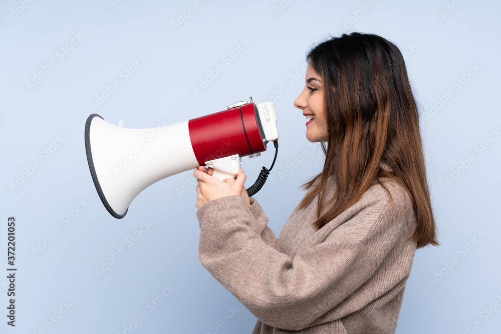 Young brunette woman wearing a sweater over isolated blue background shouting through a megaphone Young brunette woman wearing a sweater over isolated blue background shouting through a megaphone