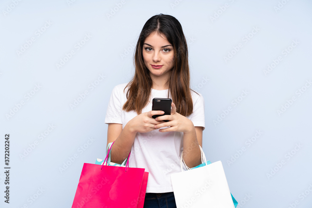 Young brunette woman over isolated blue background holding shopping bags and writing a message with her cell phone to a friend Young brunette woman over isolated blue background holding shopping bags and writing a message with her cell phone to a friend