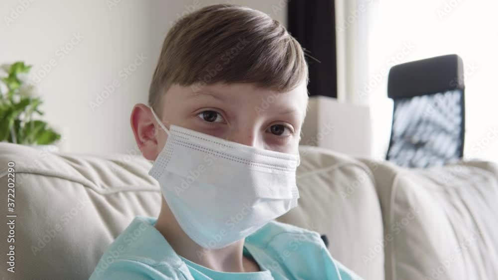 A young boy in a face mask looks at the camera as he sits on a couch at home - face closeup A young boy in a face mask looks at the camera as he sits on a couch at home - face closeup