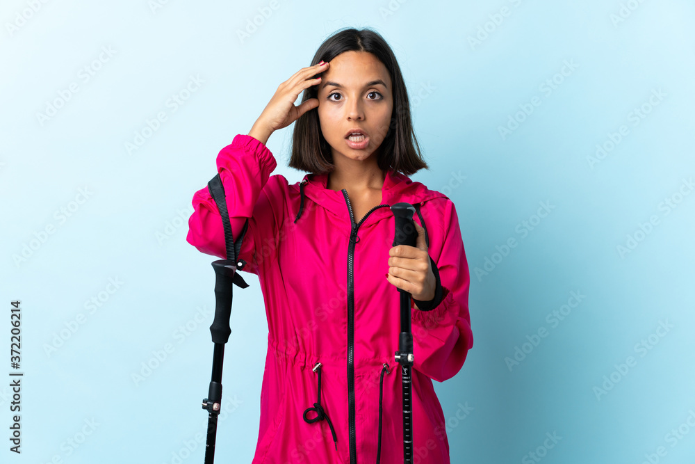 Young latin woman with backpack and trekking poles isolated on blue background with surprise expression