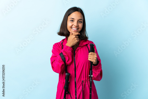 Young latin woman with backpack and trekking poles isolated on blue background happy and smiling © luismolinero