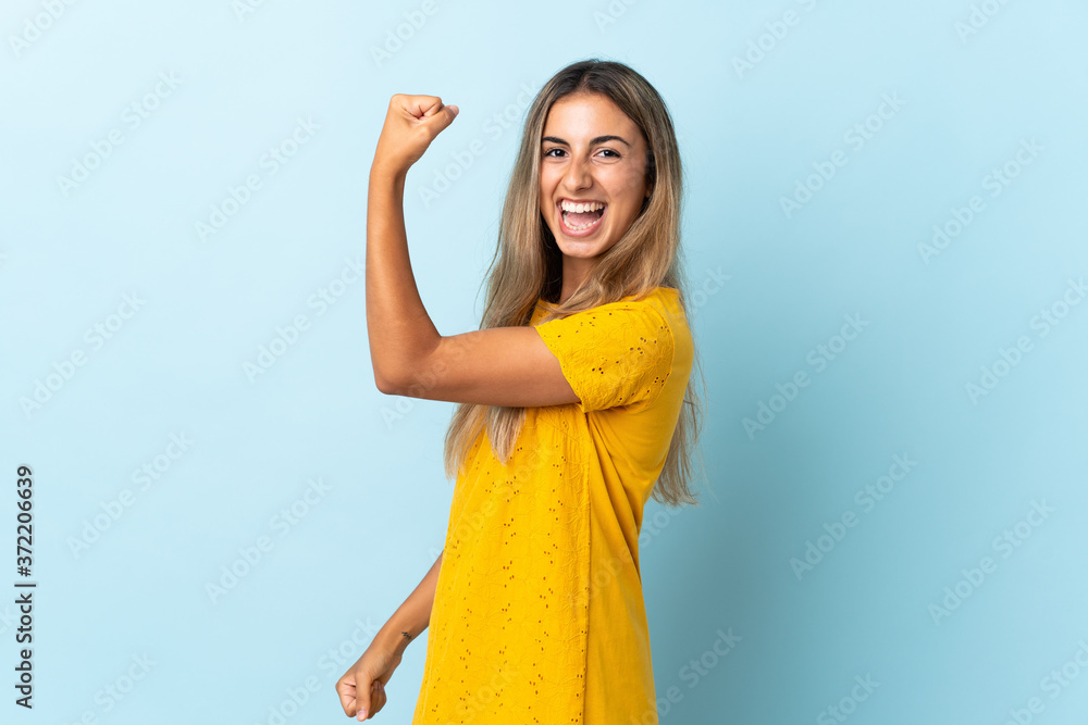 Young hispanic woman over isolated blue background doing strong gesture Young hispanic woman over isolated blue background doing strong gesture