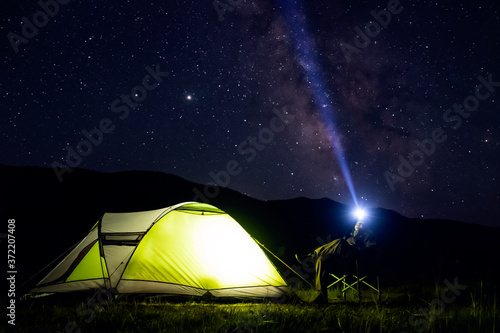 Illuminated green tent with man sitting near and mountains silhouette and many stars background. Hiking, tourism concept. Adventure travel. Outdoor landscape. Night sky, stars background.