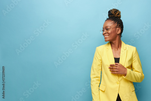 portrait photo of dark-skinned beautiful girl on a blue background in a yellow jacket, transparent glasses she looks away and smiles
