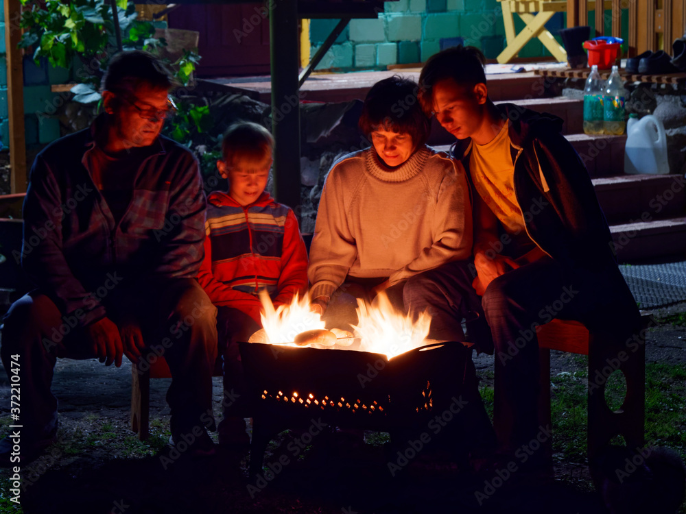 Group of young friends sitting by the fire late at night, grilling sausages and having fun
