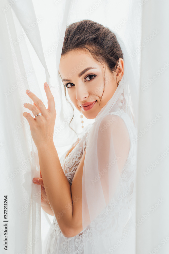 Side view of young brunette bride looking at camera while touching curtains at home Side view of young brunette bride looking at camera while touching curtains at home