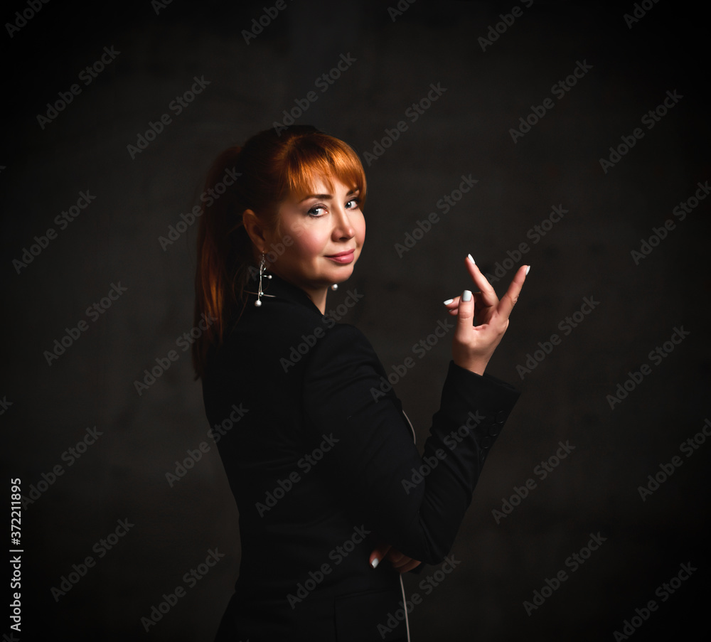 Red-haired female in jewelry and black pantsuit. She smiling and showing her forefinger, looking back. Posing on gray background Red-haired female in jewelry and black pantsuit. She smiling and showing her forefinger, looking back. Posing on gray background
