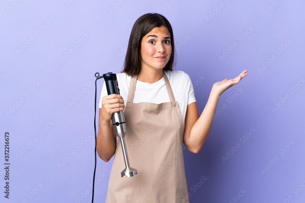 Brazilian woman using hand blender isolated on purple background having doubts while raising hands Brazilian woman using hand blender isolated on purple background having doubts while raising hands