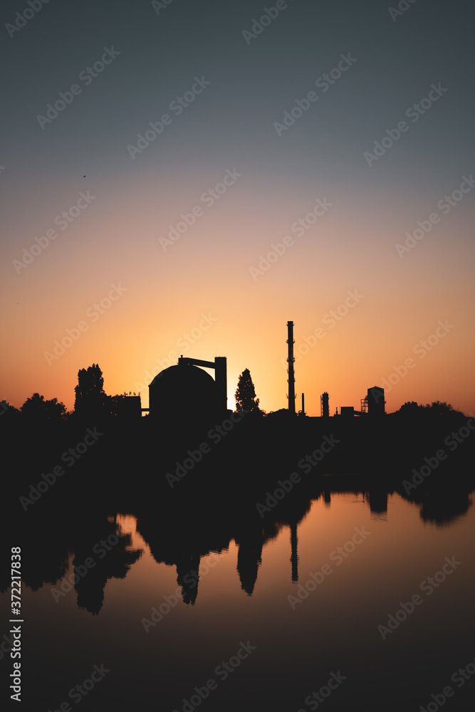Production facility silo at sunset.  Industrial complex located by the lake. Grain or other loose product storehouse silhouette on the horizon. Water reflection. 