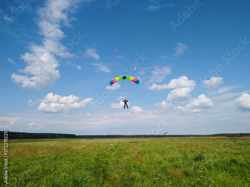 A skydiver with a bright multicolored parachute flies against the background of a blue sky with white sparse clouds and green grass in summer