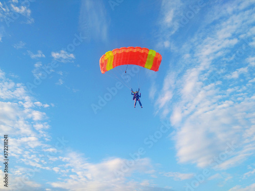 A skydiver with a bright orange parachute flies against a blue sky with white sparse clouds