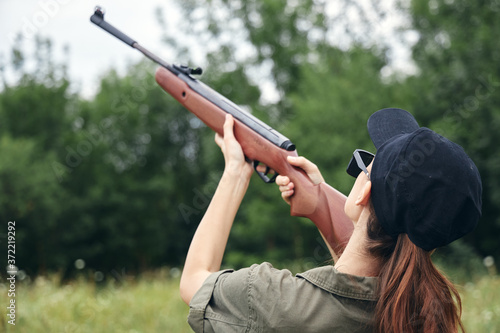 Woman on nature aim up gun hunting back view black cap fresh air green 