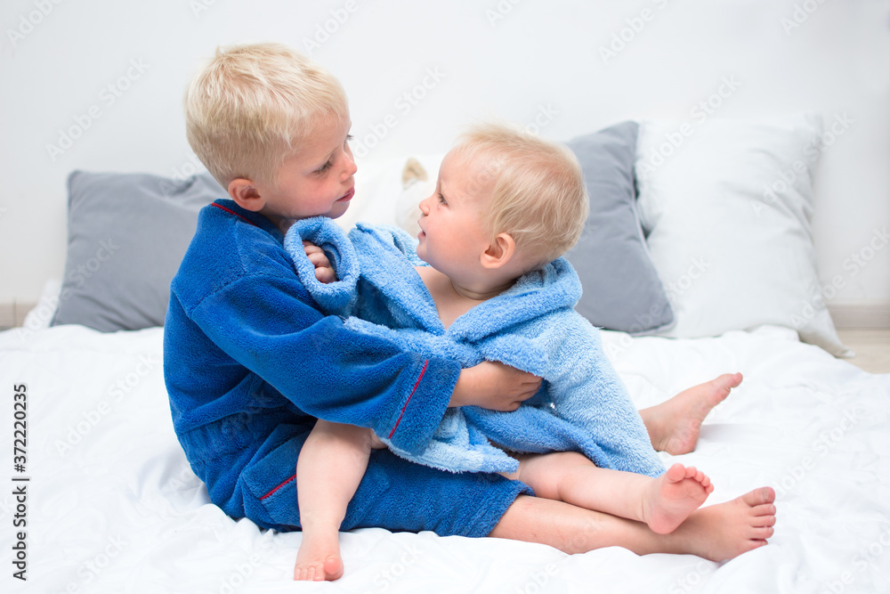 Baby brothers sitting on bed in bathrobe and playing. Siblings in blue bathrobe. Baby care concept, banner copy space Baby brothers sitting on bed in bathrobe and playing. Siblings in blue bathrobe. Baby care concept, banner copy space