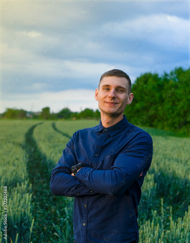 A young guy in a shirt stands in a green field, looks at the camera and crosses his arms over his chest at sunset.