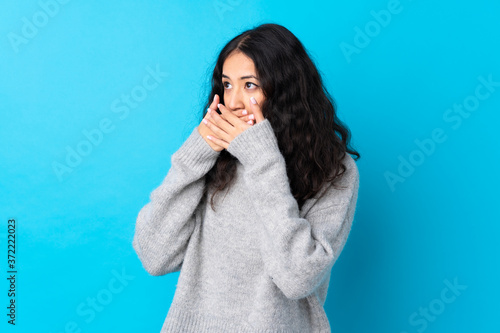 Spanish Chinese woman over isolated blue background covering mouth and looking to the side
