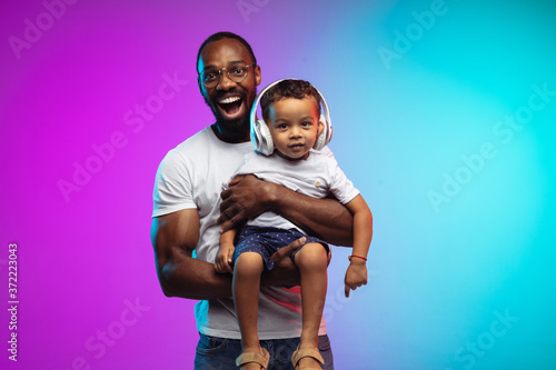 African-american father and son portrait on gradient studio background in neon. Beautiful male models in casual style, white shirt. Concept of human emotions, facial expression, sales, ad, family. photo