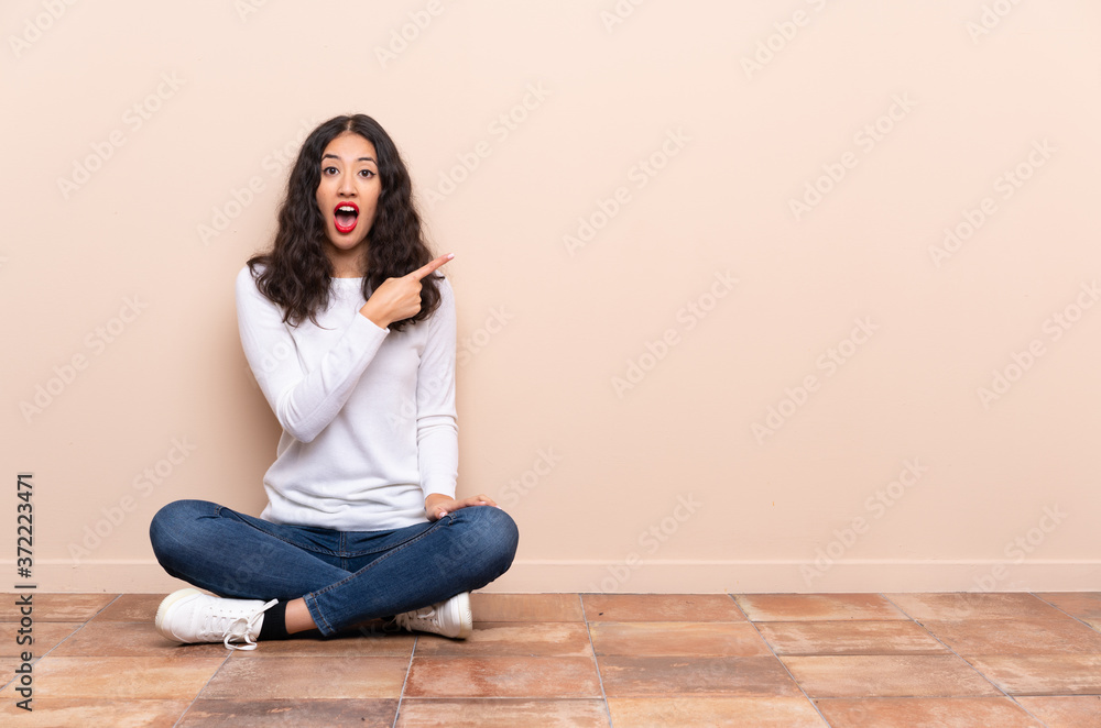 Young woman sitting on the floor surprised and pointing side