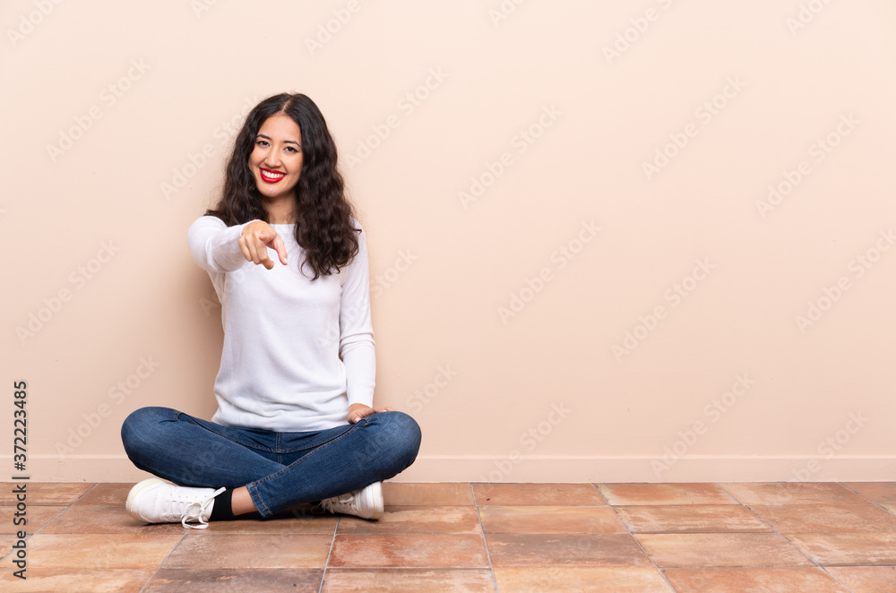 Young woman sitting on the floor points finger at you with a confident expression